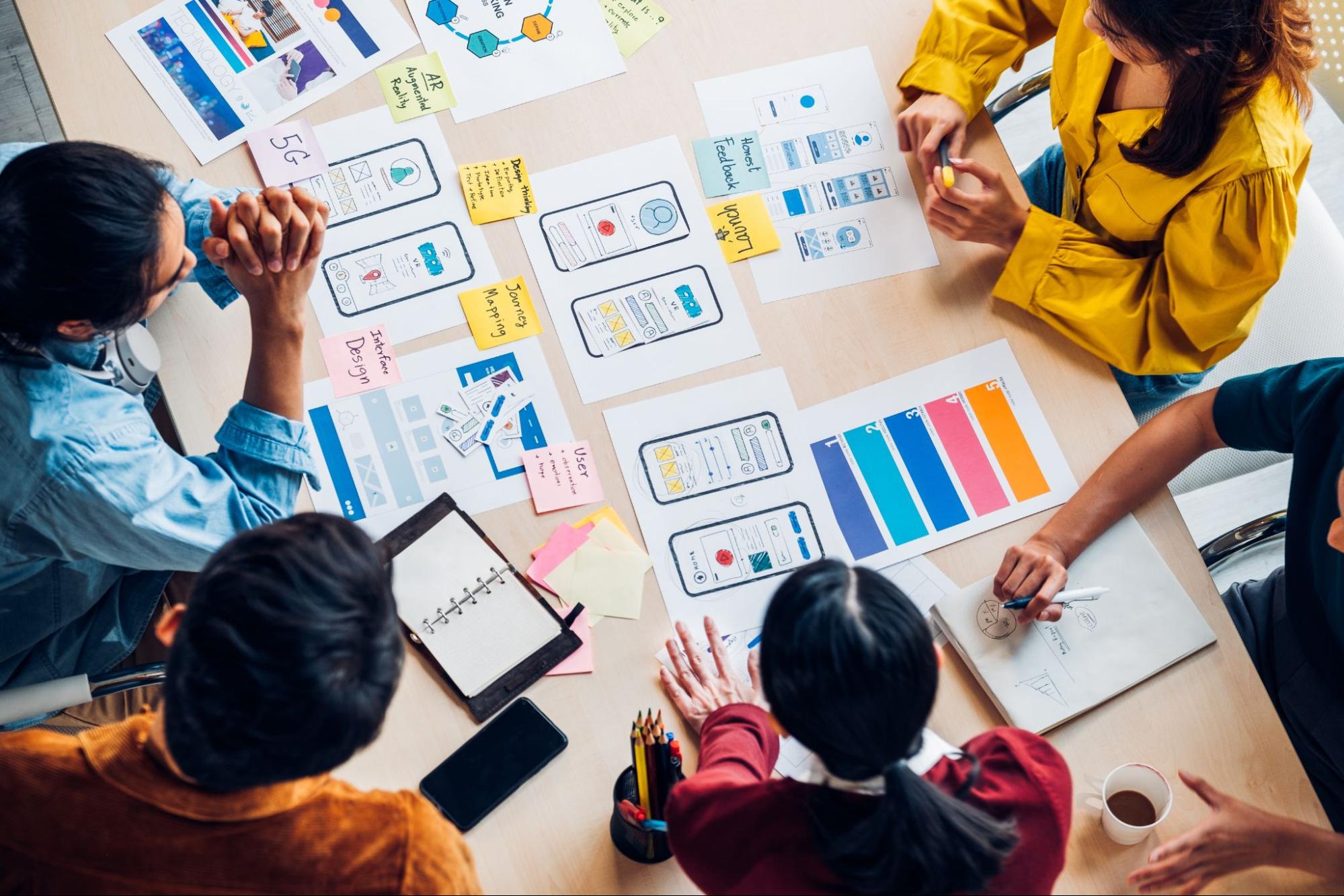 Stock photo showing an overhead view of five co-workers collaborating around a table and looking at sketches of a mobile app