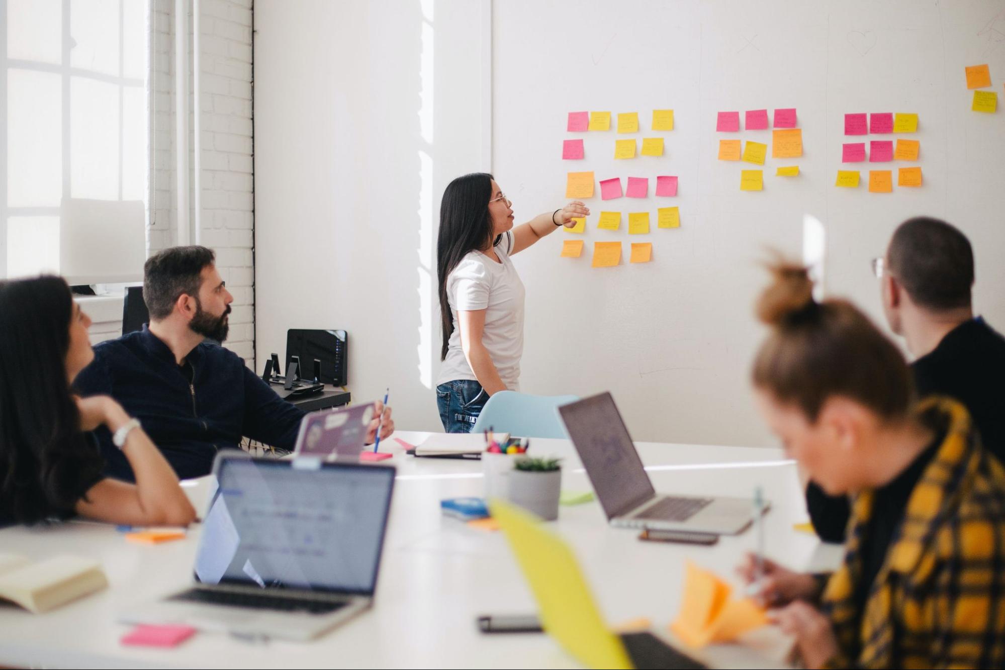 Stock photo of a person discussing sticky notes on a whiteboard with a table of teammates looking on