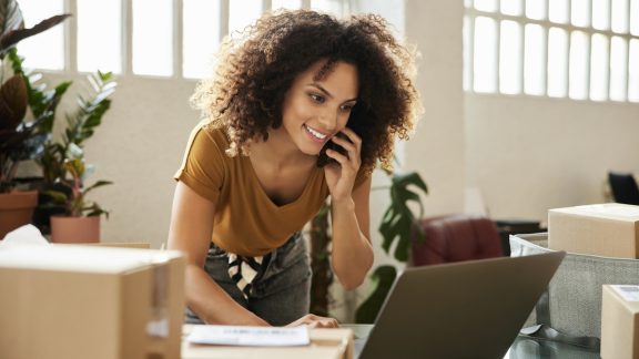 Young female talking on smart phone while using laptop. Confident businesswoman is working at home office. She is having curly hair.