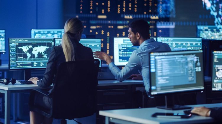 Confident Female Data Scientist Works Closely to her Young Colleague in Big Control and Monitoring Room with Neural Network. Young Specialists Talk and Discuss Work in a Room with Other Employees.