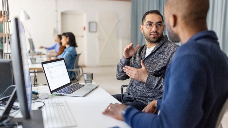 Two young male application developers discussing certain codes for an application while sitting at coworking office desk. Software developers planning how to create innovative software.
