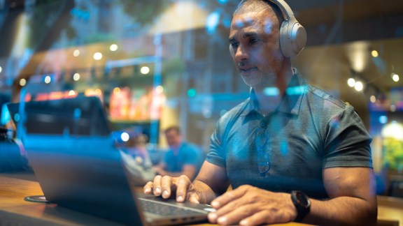 A man wearing headphones focuses on his laptop while seated in a modern cafe.