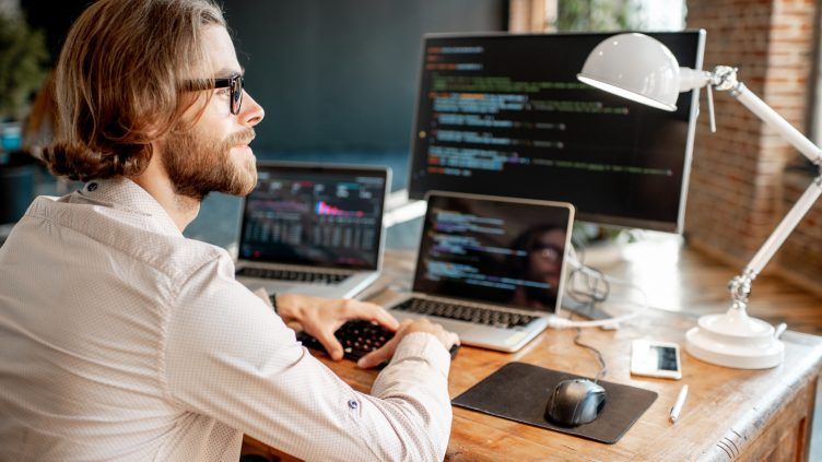 Young male programmer writing a program code sitting at the workplace with three monitors in the office