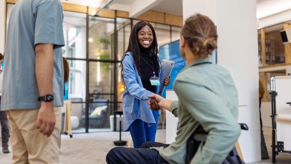 African woman shaking hands with businessman sitting on wheelchair at a conference. Young female professional meeting a colleague sitting in wheelchair at business conference.