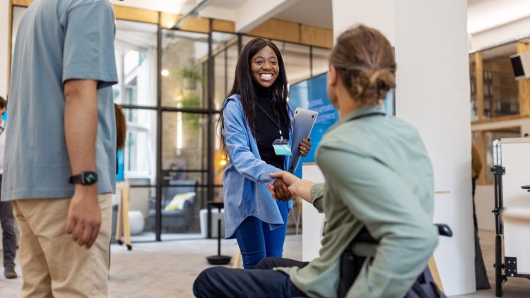 African woman shaking hands with businessman sitting on wheelchair at a conference. Young female professional meeting a colleague sitting in wheelchair at business conference.