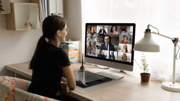 Back rear view happy millennial female employee looking at computer monitor, holding online video call negotiations meeting with diverse colleagues and male executive manager, working from home.