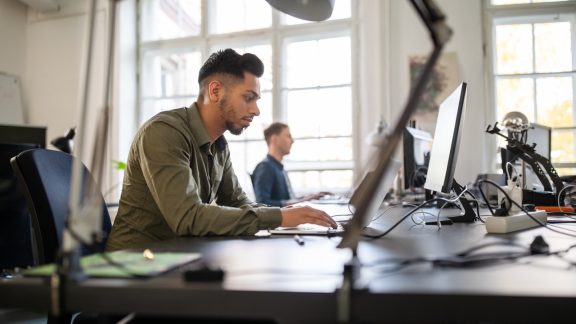 Young man busy working on laptop in office with a colleague in background. Businessman using a laptop computer at his desk.