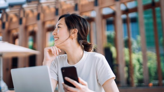 Confident young Asian freelance businesswoman looking away with smile while using smartphone and working on laptop on the go outdoors in urban park in the city. Remote working concept with flexible lifestyle