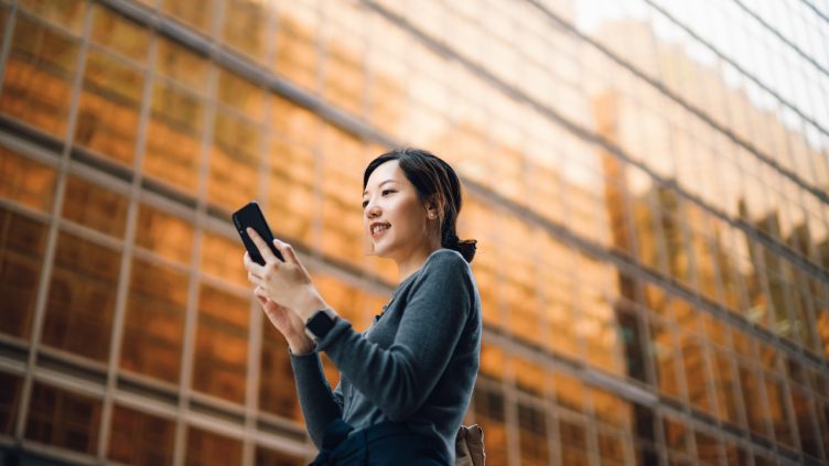 Low angle portrait of young Asian businesswoman managing online banking with mobile app on smartphone on the go. Transferring money, paying bills, checking balance in the city against corporate skyscrapers. Making business connections throughout the city