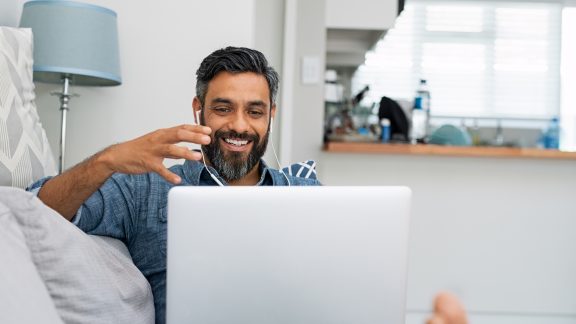 Happy mature man relaxing on couch while video calling using laptop at home. Latin man sitting on sofa and making a video call. Smiling middle eastern businessman doing online video chat while gesturing with hands.