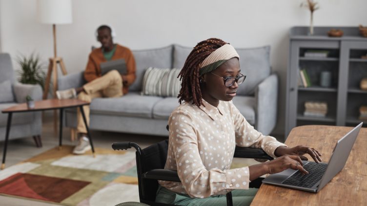 Portrait of young African-American woman using wheelchair while working from home in minimal grey interior, copy space