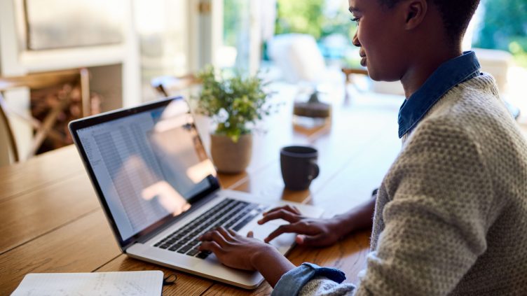 Smiling young African American female entrepreneur sitting at her dining room table at home working on a laptop