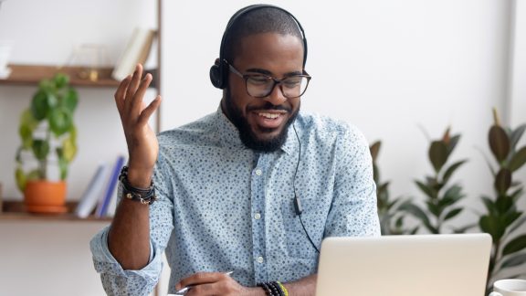 Smiling African American employee in headphones using laptop, looking at screen, making video call or watching webinar, writing notes, distance learning language concept, call center operator working