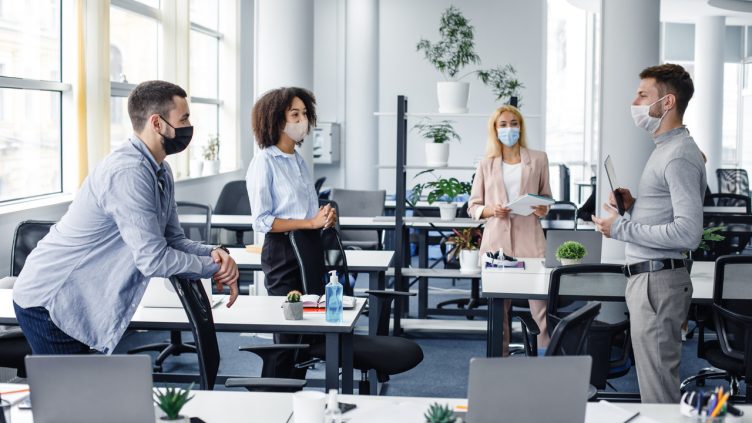 Modern team meeting, group work and social distancing. Manager with tablet speaks with workers in protective masks in interior of modern office with gadgets during coronavirus epidemic, free space