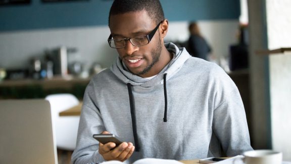 African American millennial student look at smartphone studying in cafÃ©, focused black young guy text using cellphone sitting in coffesshop working at laptop, afro busy with devices having coffee out