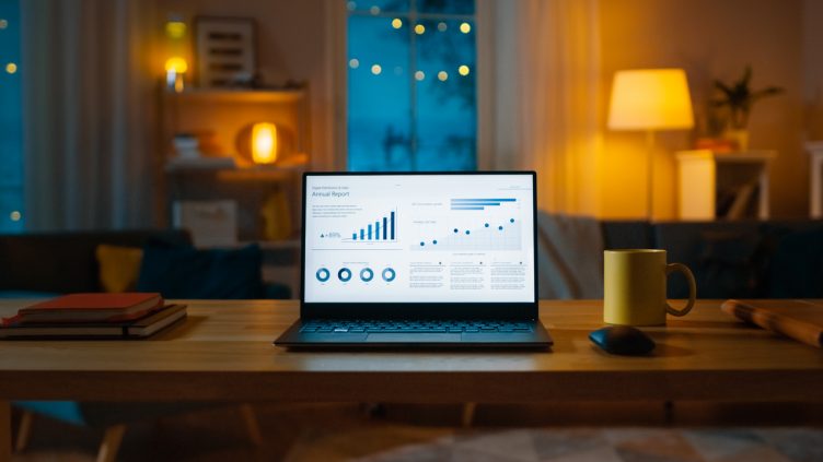 Laptop Computer Showing Statistical Infographics Stands on a Desk in the Living Room. In the Background Cozy Living Room with Warm Lights on.