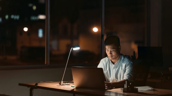 Young Asian businessman working on a laptop at his office desk at night with city lights in the background