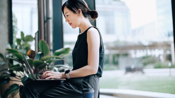 Confident and modern young Asian businesswoman working on laptop in a contemporary office space, sitting next to green plants by the window. She is wearing smart casuals in office. Female leadership. Business on the go