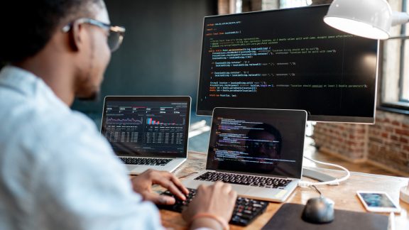 Young african male programmer writing program code sitting at the workplace with three monitors in the office. Image focused on the screen