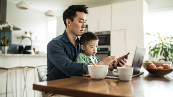 Father multi-tasking with young son (2 yrs) at kitchen table