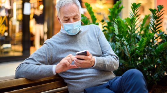 Senior man sitting on a bench and using a smartphone in a mall wearing mask, coronavirus concept