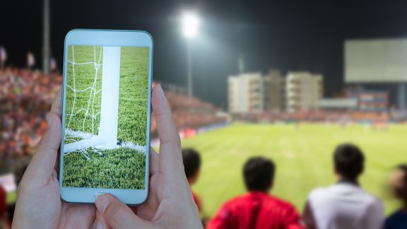 Hand holding smartphone with soccer screen, blurred football stadium background.