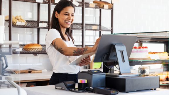 Happy woman working in bakery, pastry shop using tablet and cash computer