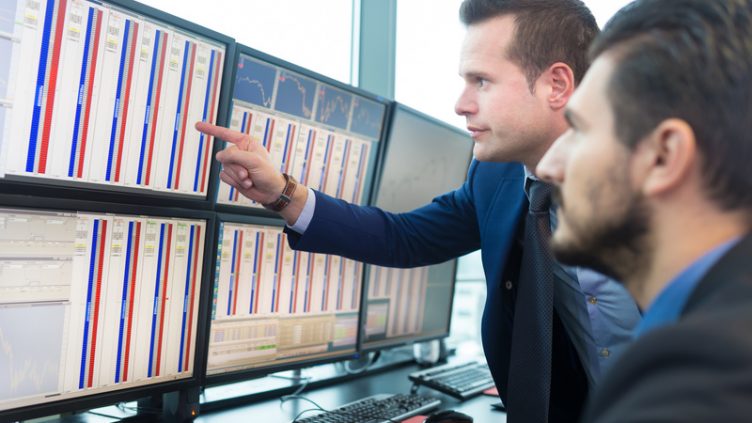 Businessmen trading stocks. Stock traders looking at graphs, indexes and numbers on multiple computer screens. Colleagues in discussion in traders office.