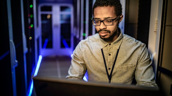 Front view of an IT engineer of African ethnicity sitting in a server room and using his laptop.