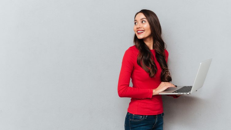 Side view of smiling brunette woman in red blouse holding laptop computer and looking back over gray background