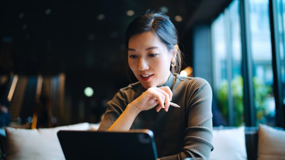 Young Asian businesswoman looking at her digital tablet while sitting at a desk in stylish co-working space and working. Successful female entrepreneur and leadership. Technology in a modern lifestyle. Working remotely with flexibility